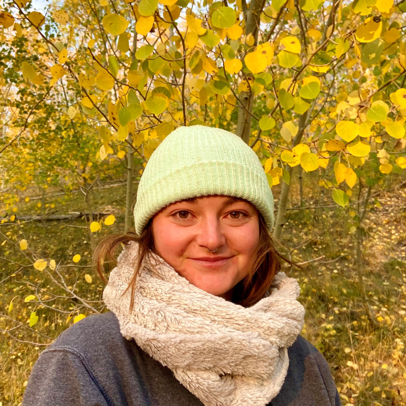 A woman with fair skin and reddish-brown hair smiles at the camera. She is wearing a light green beanie, a gray sweatshirt, and a cream-colored scarf. The background features trees with bright yellow leaves, suggesting it is autumn. The lighting is soft and natural, highlighting the colors of the fall foliage.
